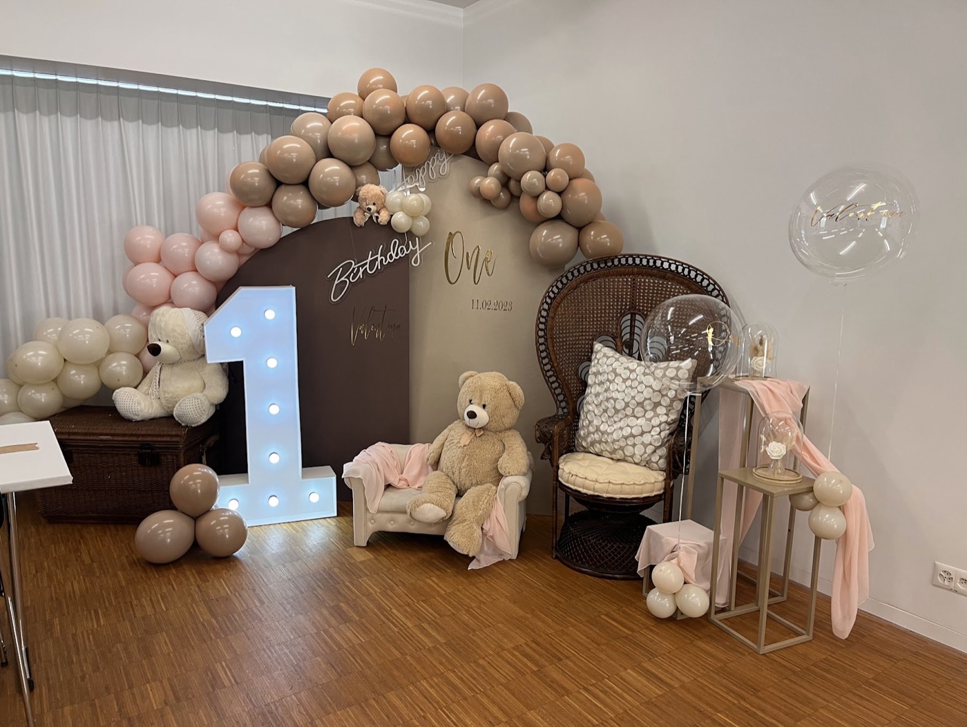 Leuchtzahl 1 vor einem Ballonbogen in Beige mit Rattan-Sessel und Teddybär